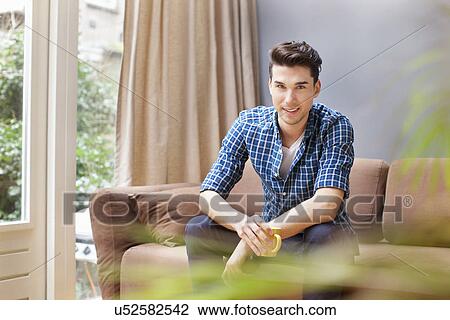 Stock Image - Portrait of smiling young man on sofa with coffee cup. Fotosearch