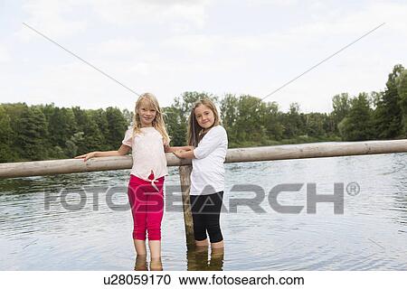 Stock Image - Portrait of two girls holding onto fence whilst paddling in rural lake. Fotosearch