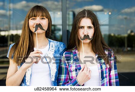 Picture - Portrait of two young women holding up mustache costume masks. Fotosearch