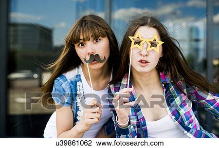Stock Image - Portrait of two young women holding up mustache and specs costume masks. Fotosearch