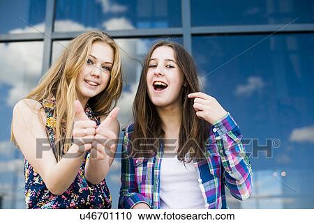 Stock Image - Portrait of two young women making thumbs up in front of glass fronted office. Fotosearch
