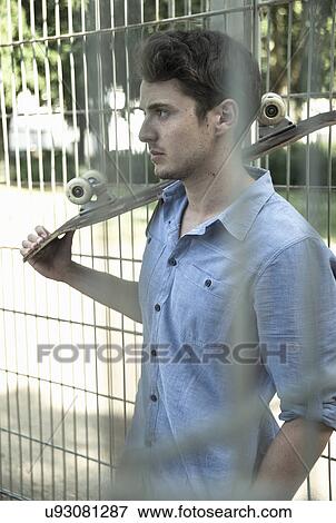 Serious young man with skateboard on shoulder behind park fence View Large Photo Image Stock Photo - Serious young man with skateboard on shoulder behind park fence. Fotosearch