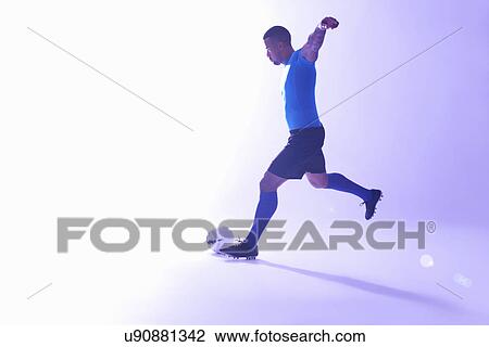 Stock Image - Studio shot of young male soccer player with arms outstretched kicking the ball. Fotosearch