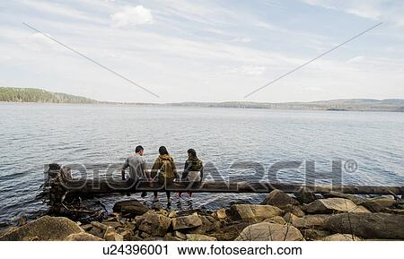 Stock Image - Three hiking friends sitting in a row on fallen tree at lakeside. Fotosearch