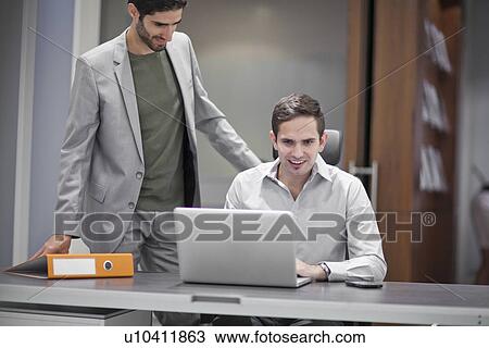 Stock Image - Two men in office, one using laptop. Fotosearch