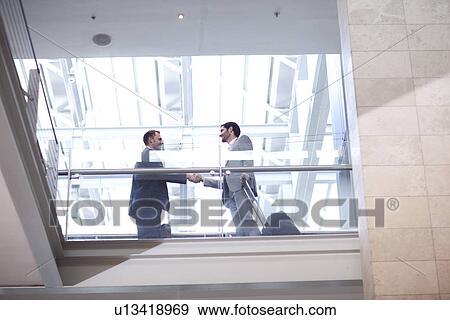 Stock Photo - Two young businessmen shaking hands on conference centre balcony. Fotosearch