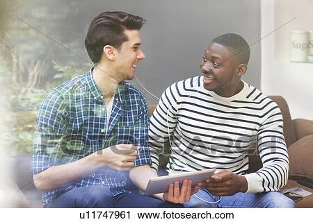 Stock Image - Two young male friends listening to music on digital tablet behind window. Fotosearch
