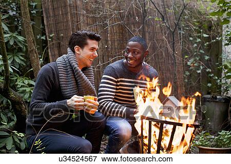 Stock Photo - Two young men crouching in front of garden fire with coffee. Fotosearch