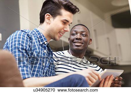 Stock Image - Two young men listening to music on digital tablet on living room sofa. Fotosearch