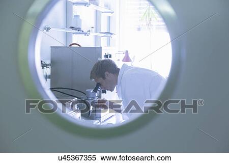 Stock Photography - View through door porthole of male scientist using microscope in lab. Fotosearch