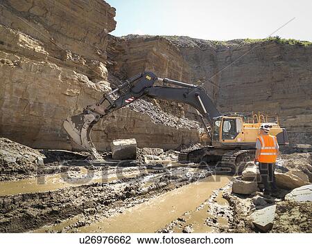 Stock Image - Worker standing next to excavator inspecting rock face in quarry. Fotosearch