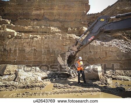 Stock Photo - Worker standing next to excavator inspecting stone in quarry. Fotosearch