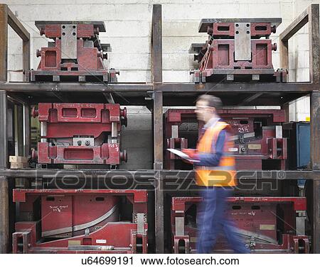 Stock Image - Worker with clipboard walking past press moulds in sheet metal factory. Fotosearch