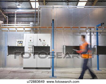 Stock Photo - Worker with clipboard walking past paint spray machine in sheet metal factory. Fotosearch