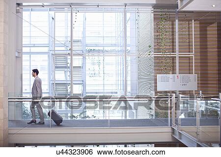Stock Photograph - Young businessman with wheeled case in conference centre. Fotosearch