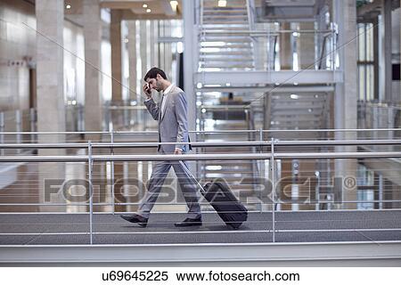 Stock Photography - Young businessman with wheeled case on conference centre walkway. Fotosearch
