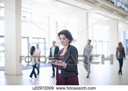 Stock Photo - Young businesswoman texting on smartphone in conference centre corridor. Fotosearch