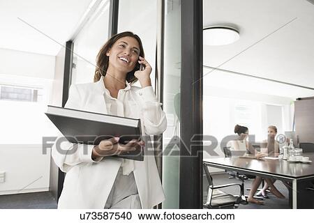 Stock Image - Young businesswoman with portfolio chatting on smartphone. Fotosearch