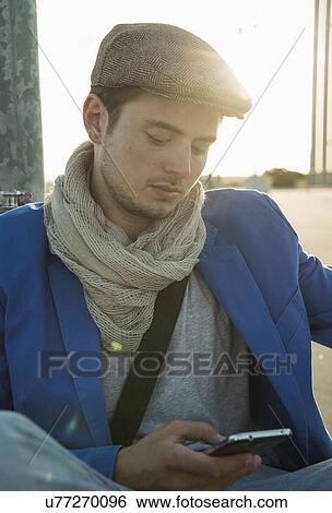 Stock Photograph - Young man leaning against wall texting on smartphone. Fotosearch
