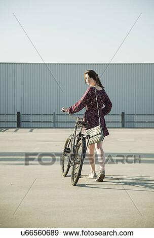 Young woman looking back whilst pushing bicycle in empty parking lot View Large Photo Image Stock Photo - Young woman looking back whilst pushing bicycle in empty parking lot. Fotosearch