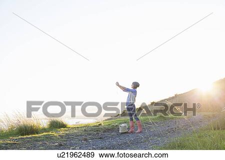Stock Photo - Young woman taking photograph on hill at sunset. Fotosearch