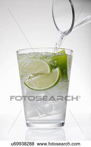 Stock Photo - Clear water pouring from jug into drinking glass with lime and ice cubes. Fotosearch