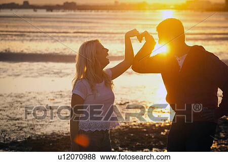 Couple forming heart shape with arms on beach at sunset View Large Photo Image Stock Photo - Couple forming heart shape with arms on beach at sunset. Fotosearch