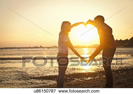 Couple forming heart shape with arms on beach at sunset View Large Photo Image Stock Photo - Couple forming heart shape with arms on beach at sunset. Fotosearch
