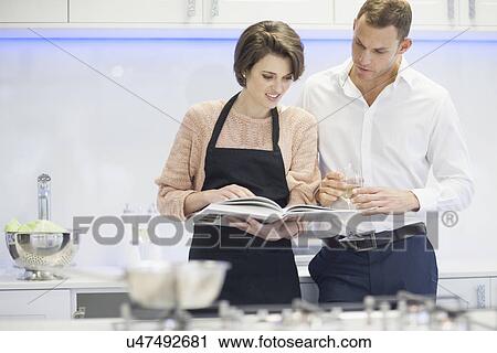 Stock Image - Couple looking at cook book and drinking glass of white wine in kitchen. Fotosearch
