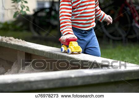 Stock Image - Cropped shot of male toddler pushing toy car around sand pit in garden. Fotosearch