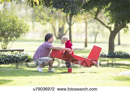 Stock Image - Father crouching to prepare toy airplane for his son in park. Fotosearch