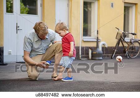 Stock Image - Father giving toddler son a hand to put on trainer in garden. Fotosearch