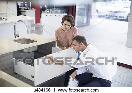 Stock Image - Female customer and salesman looking at drawer in kitchen showroom. Fotosearch