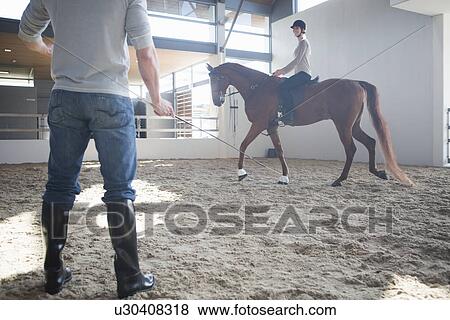 Stock Photo - Female horseback riding with instructor in indoor paddock. Fotosearch