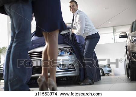 Picture - Low angle view of salesman uncovering new car for mid adult couple in car dealership. Fotosearch