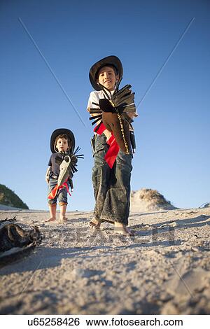 Stock Photograph - Low angle view of two brothers dressed as cowboys with hobby horses in sand dunes. Fotosearch