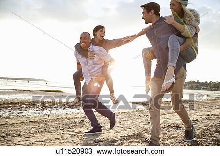 Stock Image - Men giving piggyback ride to women on beach. Fotosearch