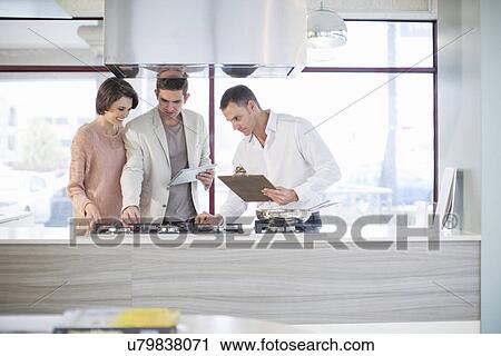 Stock Image - Mid adult couple and salesman looking at hob in kitchen showroom. Fotosearch