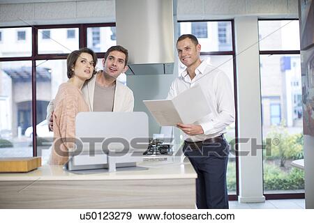 Stock Photo - Mid adult couple and salesman looking up in kitchen showroom. Fotosearch