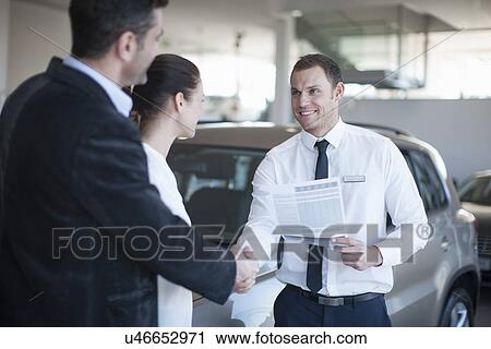 Stock Image - Mid adult couple making deal with salesman in car dealership. Fotosearch