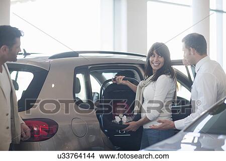 Picture - Mid adult couple with baby girl trying out baby seat in car dealership. Fotosearch