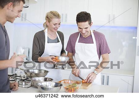 Stock Image - Mid adult man chopping carrots whilst friends watch in kitchen. Fotosearch