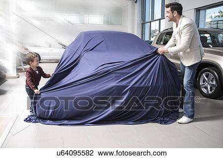 Stock Image - Mid adult man uncovering new car with son in car dealership. Fotosearch