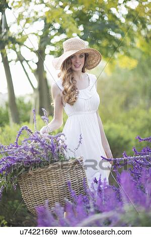 Stock Photo - Portrait of beautiful young woman in meadow carrying basket of purple flowers. Fotosearch
