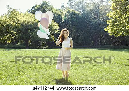 Picture - Portrait of shy young woman in park holding a bunch of balloons. Fotosearch