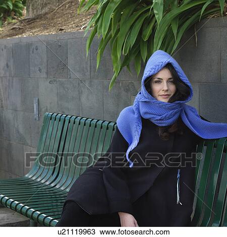 Stock Image - Portrait of young woman in snood scarf sitting on bench. Fotosearch
