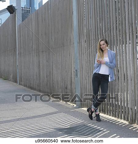 Portrait of young woman with smartphone leaning against fence View Large Photo Image Stock Image - Portrait of young woman with smartphone leaning against fence. Fotosearch