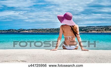Stock Photograph - Rear view of young woman wearing bikini and sunhat on beach, La Maddalena, Sardinia, Italy. Fotosearch