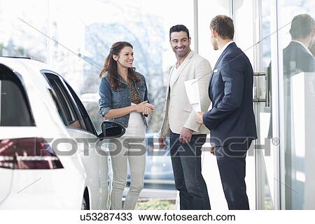 Stock Image - Salesman and mid adult couple chatting in car dealership. Fotosearch