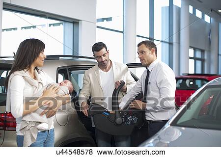 Stock Photo - Salesman chatting to mid adult couple with baby girl in car dealership. Fotosearch